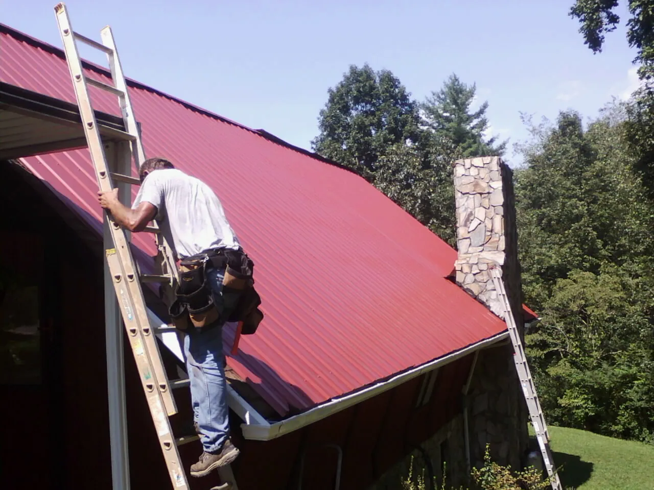 Roofer on a ladder beside a red metal roof and stone chimney — Soffit & Fascia Repair by Roofing Brothers in Mccordsville